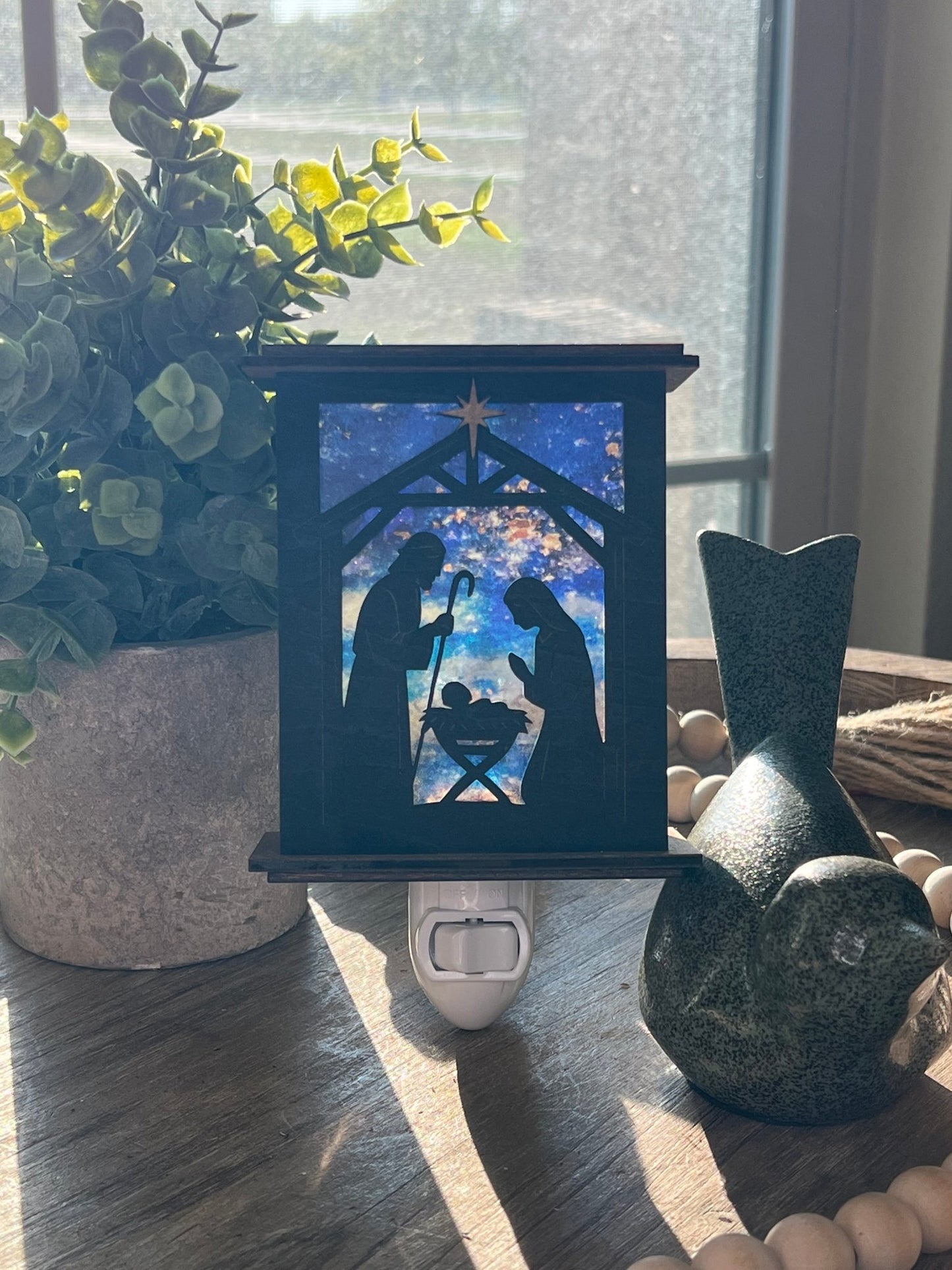 Rustic nativity nightlight lantern made of black wood and golden oak stained acrylic standingup with front view and sunlit.